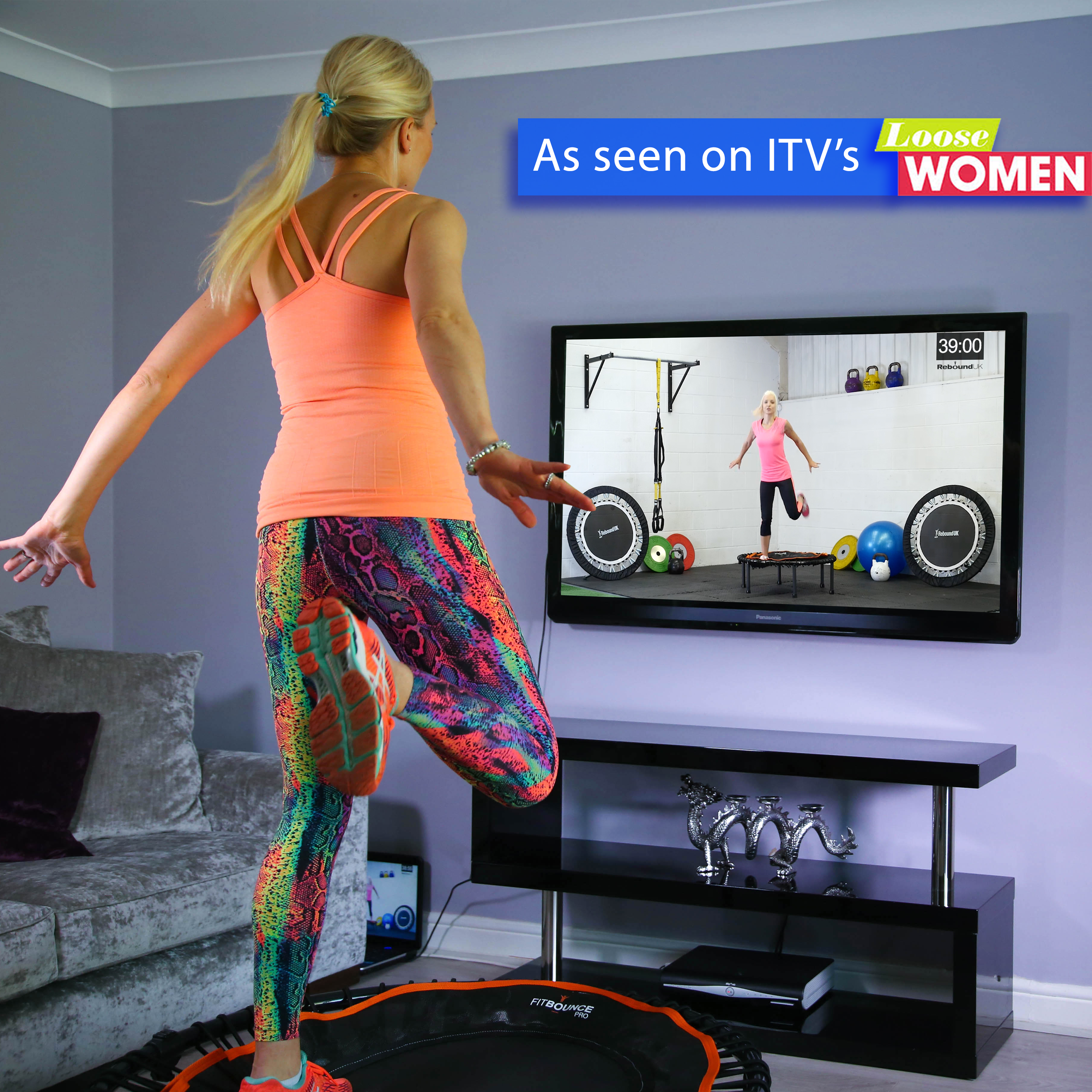 Woman bouncing in living room on fit bounce pro rebounder watching online workout on TV from Rebound Fitness